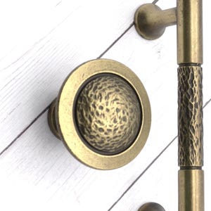 May include: Antique brass door hardware set, including a round knob with a textured center and a long pull handle with a textured center section. The hardware is set against a white wood background.