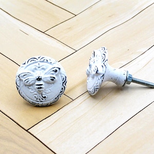 May include: Two white, distressed-finish cabinet knobs with a bee motif. One knob is upright, the other is on its side. The knobs have a round shape with a detailed bee design and a decorative edge. The hardware is silver.