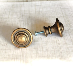 May include: Two antique-style bronze cabinet knobs with a screw. One knob is round with concentric circles, the other is a decorative shape. The knobs have a distressed finish, suggesting age or vintage style. They are likely for furniture restoration or adding a classic touch.