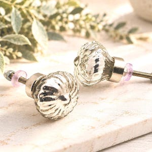 May include: Two silver-toned, pumpkin-shaped cabinet knobs with a reflective, metallic finish. Each knob features a ribbed, textured surface, a short metal stem, a small translucent pink washer, a threaded bolt, and a hexagonal nut at the end.