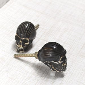 May include: Two decorative skull cabinet knobs. The knobs are black with off-white accents, featuring detailed skull faces. Each knob has a threaded metal post for installation, perfect for adding a unique touch to furniture or decor.