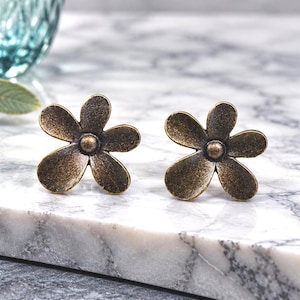 May include: A pair of stud earrings shaped like five-petaled flowers. The earrings are made of a dark, antiqued bronze-toned metal with a textured, matte finish and a small raised circular center point on each flower.