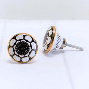May include: Two decorative cabinet knobs. Each knob features a white and black floral design with a gold-colored rim. The knobs have a screw for easy installation. The knobs are perfect for adding a touch of elegance to any cabinet or drawer.