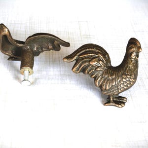 May include: Two bronze-colored rooster-shaped cabinet pulls. One pull is fully assembled with a screw, while the other is a complete rooster figure. The pulls are detailed with feathers and a comb.
