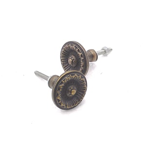May include: Two antique bronze cabinet knobs with a detailed, circular design. Each knob has a central raised circle surrounded by a textured pattern. The knobs are attached to silver-colored screws, ready for installation. The hardware has a vintage aesthetic.