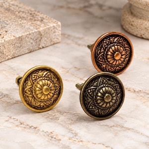 May include: Three round metal cabinet knobs featuring a raised floral mandala pattern. One knob is finished in a bright gold tone, one in a dark bronze tone, and one in a copper tone. Each knob has a central circular dome and a threaded bolt.