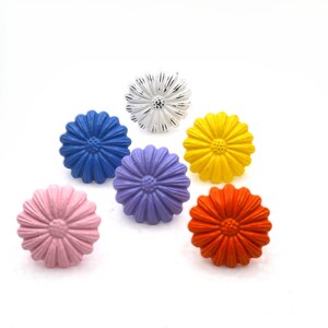 May include: A collection of six daisy-shaped cabinet knobs in various colors. The knobs are painted in shades of pink, blue, purple, yellow, and orange, with a distressed white knob. These decorative knobs are ideal for adding a pop of color to furniture.