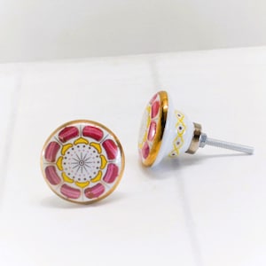 May include: Two decorative cabinet knobs. The knobs are white with a gold rim and feature a floral design in pink and yellow. The knobs have a metal screw for installation.