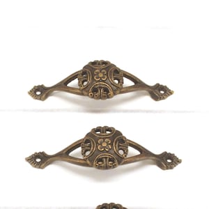May include: Ornate bronze-toned drawer pulls with a floral design. The pulls have a curved shape with decorative detailing and are mounted on a white surface. These vintage-style pulls are suitable for furniture restoration projects.