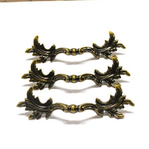 May include: Three antique bronze drawer pulls with ornate, symmetrical leaf and scroll designs. The hardware is made of metal and has a vintage aesthetic. The pulls are arranged in a horizontal stack against a white background.
