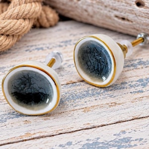 May include: Two round cabinet knobs featuring a concave center with a dark gray crackle glaze finish. Each knob has a smooth white ceramic border, a thin gold-toned metal rim, and a gold-toned metal screw post with a clear plastic washer.