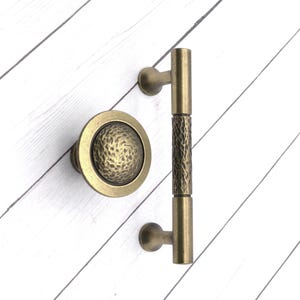 May include: Antique brass door hardware set, including a round knob with a textured center and a vertical pull handle. The handle has a textured center section. The hardware is set against a white wood background.