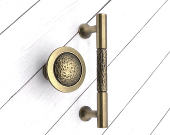 May include: Antique brass door hardware set, including a round knob with a textured center and a vertical pull handle. The handle has a textured center section. The hardware is set against a white wood background.