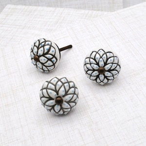 May include: Three decorative ceramic cabinet knobs. Each knob is white with a black floral design and small blue dots. The knobs have a bronze-colored base and a metal screw for installation.