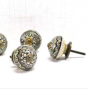 May include: Decorative ceramic cabinet knobs with a floral design in gray, white, and gold. The knobs have a round shape and a brass-colored base. The knobs are for furniture and home decor.