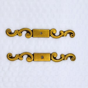 May include: Two antique brass drawer pulls with a decorative scroll design. Each pull has a rectangular center with a single screw hole and ornate, curved ends. The pulls are set against a white background.