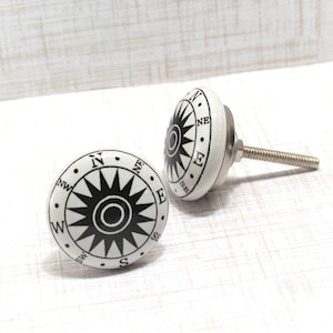 May include: Two white ceramic cabinet knobs with a black compass rose design. The knobs have a silver metal screw and are set against a white wooden background. The compass rose includes the cardinal directions.