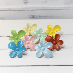 May include: A collection of colorful flower-shaped decorative accents in various shades, including blue, green, yellow, pink, and red. These small, glossy floral embellishments are arranged on a white surface, offering a cheerful and vibrant aesthetic.