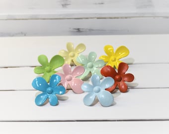 May include: A collection of colorful flower-shaped decorative accents in various shades, including blue, green, yellow, pink, and red. These small, glossy floral embellishments are arranged on a white surface, offering a cheerful and vibrant aesthetic.