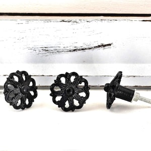 May include: Three black decorative cabinet knobs with a floral design. Two knobs are face up, and one is on its side, showing the screw. The knobs are made of metal and have a matte finish.