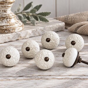 May include: A set of six round cabinet knobs featuring a cream-colored ceramic surface with an embossed swirling floral pattern and a dark bronze-toned metal center accent. One knob is shown from the side, revealing a threaded metal bolt.