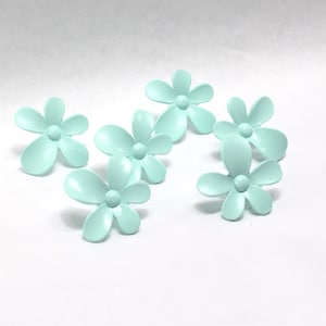 May include: Six light blue flower-shaped decorative embellishments. Each flower has five rounded petals and a small, round center. The flowers are arranged on a white surface, casting soft shadows.