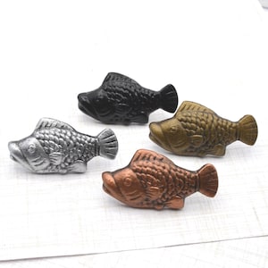 May include: Four decorative fish figurines in varying metallic finishes: black, silver, bronze, and gold. Each fish has a textured scale pattern and a detailed fin design. The figurines are arranged on a white surface.