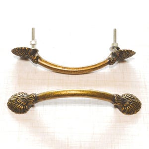 May include: Two antique-style bronze cabinet pulls. Each pull has a curved handle with decorative, shell-like ends. One pull is assembled with a bolt and nut. The pulls are set against a light-colored surface.