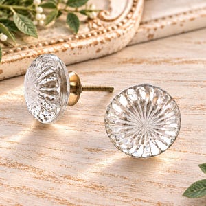 May include: Two round cabinet knobs made of clear, faceted glass with a sunburst pattern radiating from the center. Each knob features a metallic gold-toned base and a threaded metal screw for attachment to furniture or cabinetry doors.