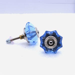 May include: Two blue glass cabinet knobs with a faceted design. The knobs have a silver-colored metal base and a threaded post with a nut and washer for installation. The knobs are a light blue color and have a starburst shape.