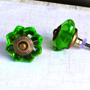May include: Two decorative cabinet knobs. The knobs feature a faceted, flower-shaped design in a vibrant green color. The base is a bronze color, and the hardware includes a screw and nut for installation.