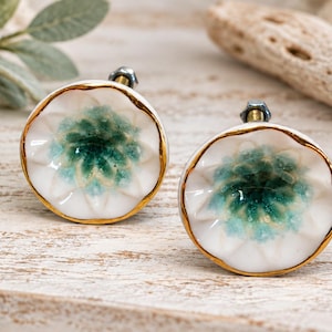 May include: Two round ceramic cabinet knobs featuring a white base with a scalloped edge and a central gradient of teal and green glaze. Each knob is finished with a metallic gold rim and includes a small metal bolt and nut attachment.