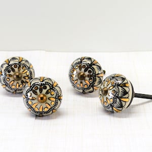 May include: Four decorative ceramic cabinet knobs. Each knob features a cream-colored base with black and gold floral patterns. The knobs have a brass-colored center and a threaded metal stem for installation. The knobs are designed for furniture or cabinet doors.