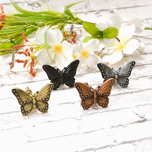 May include: Four butterfly-shaped decorative cabinet knobs in antique gold, black, bronze, and silver. The knobs are made of metal and have detailed wing patterns. The background includes white flowers and greenery.