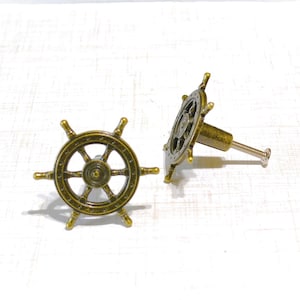 May include: Two antique brass-colored cabinet knobs in the shape of a ship's wheel. The knobs have a detailed design with spokes and a central hub. The screw is visible on the back of one knob. Nautical-themed hardware.