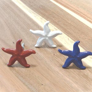 May include: Three starfish-shaped cabinet knobs in red, white, and blue. These decorative knobs are made of a solid material and are designed to add a nautical touch to furniture or decor. They are placed on a wooden surface.