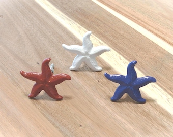 May include: Three starfish-shaped cabinet knobs in red, white, and blue. These decorative knobs are made of a solid material and are designed to add a nautical touch to furniture or decor. They are placed on a wooden surface.