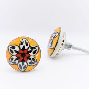May include: Two decorative ceramic cabinet knobs. Each knob features a floral design with black, white, and red petals on a yellow background. The knobs have a silver metal base and screw.