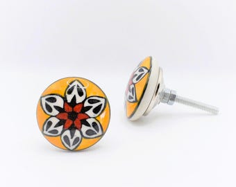 May include: Two decorative ceramic cabinet knobs. Each knob features a floral design with black, white, and red petals on a yellow background. The knobs have a silver metal base and screw.