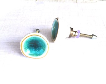 May include: Two ceramic cabinet knobs with a white base, teal center, and gold trim. One knob is detached, showing the brass hardware and screw. The knobs are designed for furniture and home decor.