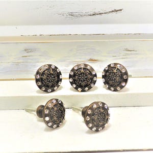 May include: Set of five bronze-colored cabinet knobs. Each knob features a decorative floral design in the center, surrounded by a ring of clear, sparkling crystals. The knobs are mounted on silver-colored screws.