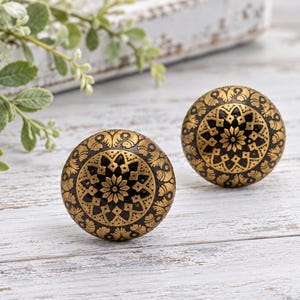 May include: Two round cabinet knobs featuring a black base with an intricate gold-colored floral and geometric pattern. The knobs have a smooth, domed surface and a metallic finish, displaying a repeating symmetrical design across the front.