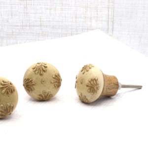 May include: Cream-colored decorative cabinet knobs with a floral design. The knobs have a wooden base and a metal screw. The floral pattern is in a darker cream color. The knobs are round and have a smooth finish.