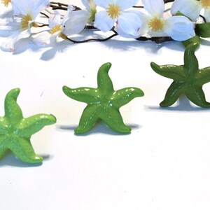 May include: Three green starfish figurines are arranged on a white surface, with white flowers in the background. The starfish have five points and a glossy finish, suggesting a decorative or craft item.