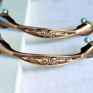 May include: Two antique-style bronze drawer pulls. Each pull features a floral design in the center and is curved with a small, light blue, spherical accent on each end. The pulls are approximately 6 inches long.