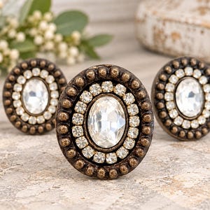 May include: Three round cabinet knobs featuring a central oval clear crystal surrounded by a ring of smaller clear rhinestones and an outer border of dark bronze metal with a beaded texture. The knobs have a metallic finish.