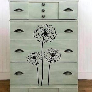 May include: A light green dresser with black hardware and a dandelion design. The dresser has five drawers and a top section with three small knobs. The dandelion design is black and covers the front of the drawers.