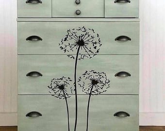 May include: A light green dresser with black hardware and a dandelion design. The dresser has five drawers and a top section with three small knobs. The dandelion design is black and covers the front of the drawers.