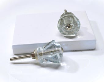 May include: Two clear glass cabinet knobs with a faceted design and silver-colored hardware. One knob is lying on a white surface, while the other is upright. The knobs are designed for furniture and home decor.