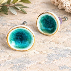 May include: Two round ceramic cabinet knobs featuring a concave center with a vibrant teal crackle glaze finish. Each knob has a white outer rim, a thin gold-colored edge, and a metal bolt with a nut attached to the back for installation.